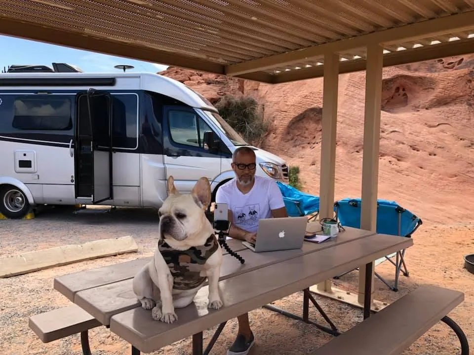 Tony working on a laptop at a picnic table, Matsu sitting on the table, the RV parked behind them among red rocks